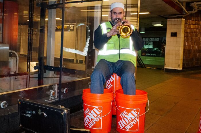 Street musician playing trumpet seated on Home Depot buckets in NYC subway capturing powerful street moments.