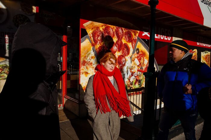 Woman in red scarf walking on NYC street with shadows and a pizza shop in the background, capturing powerful street moments
