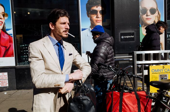 Man in beige suit smoking a cigarette on busy NYC street with vibrant advertising posters in the background, capturing street moments.