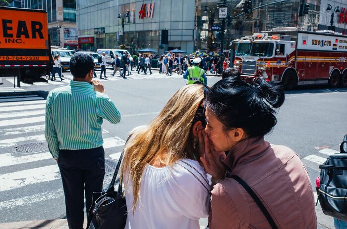 Two women sharing a secret on a busy NYC street corner amid pedestrians and a fire truck in the background.