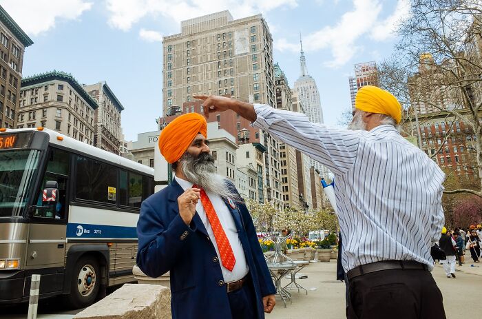 Two men wearing orange turbans engage in conversation on a busy NYC street with tall buildings and a bus nearby.