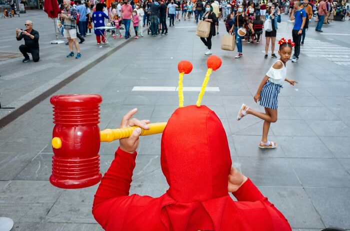 Person in a red costume holding a large toy hammer, with NYC street scenes and pedestrians in the background.