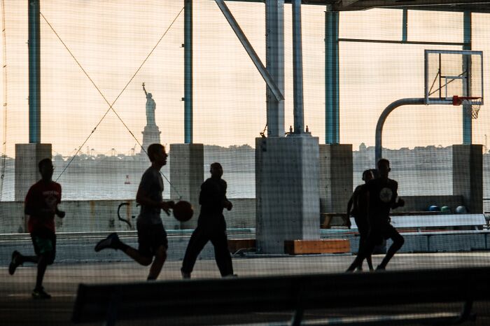 Silhouettes of people playing basketball on an outdoor court with Statue of Liberty in background, NYC street moments.