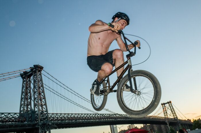 Shirtless BMX rider performing a jump in NYC street photo with bridge structure in the background at sunset.