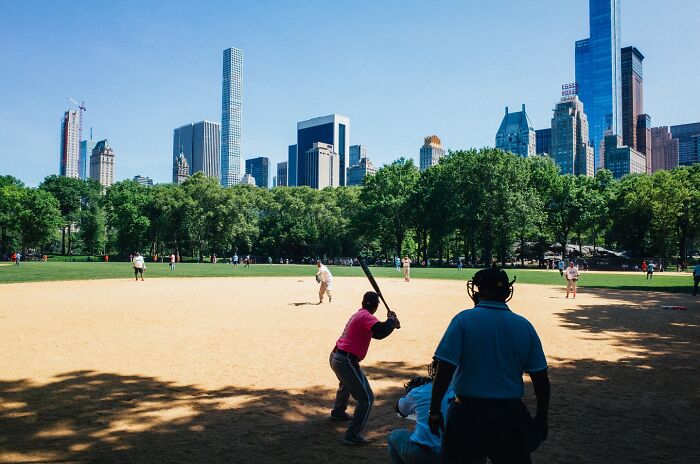 Street baseball game in NYC park with players, umpire, and city skyline in the background capturing powerful street moments.