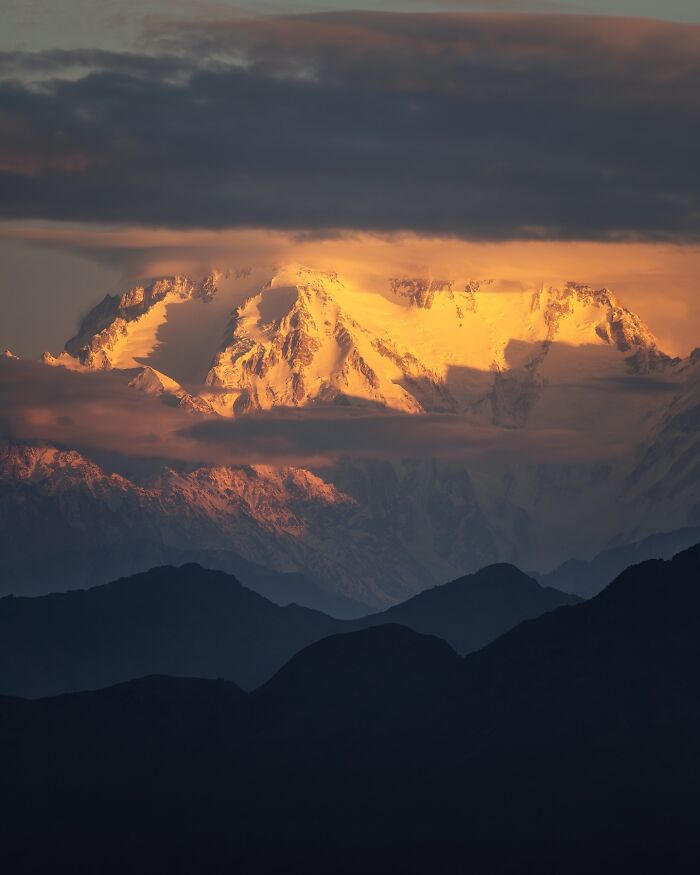 Snow-covered mountain landscape glowing at sunset with dark clouds and silhouetted hills, breathtaking landscape shot.
