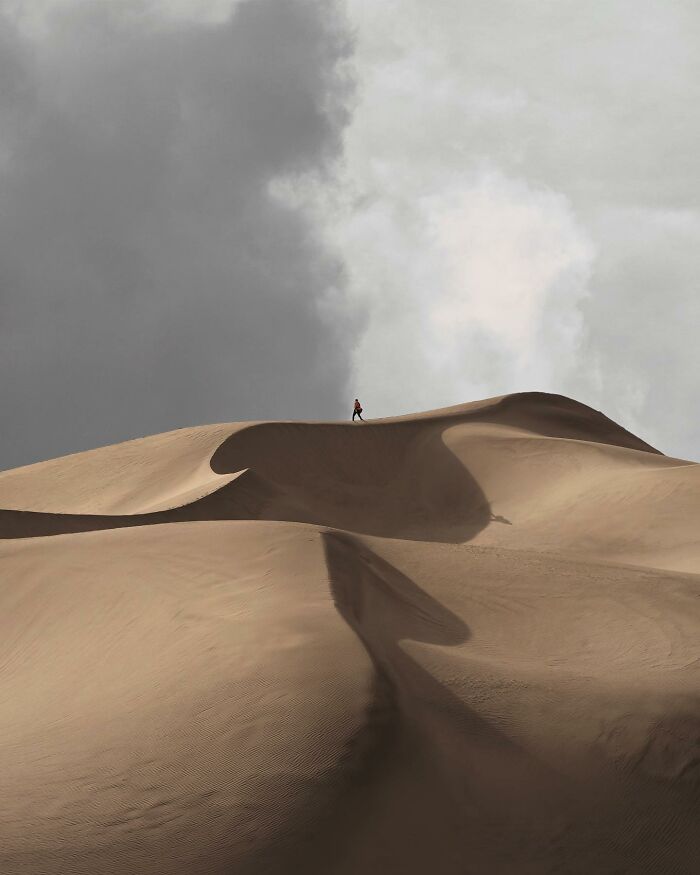 Person walking on a vast sand dune under dramatic cloudy sky in breathtaking landscape photography.