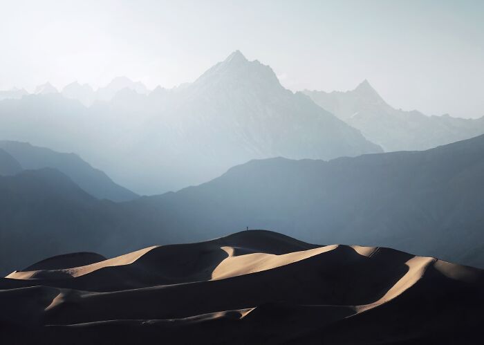 Breathtaking landscape shot of sunlit sand dunes with misty layered mountains in the background, resembling a fantasy movie scene.