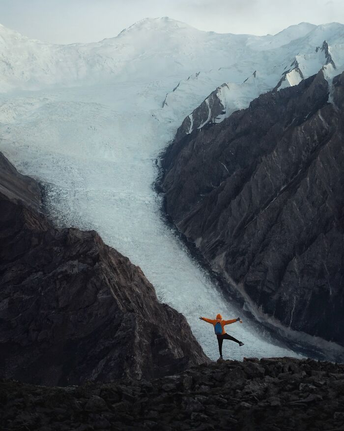Person in orange jacket posing on rocky terrain with breathtaking landscape of glaciers and mountains in the background