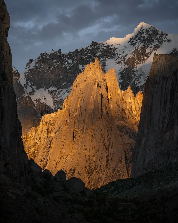 Breathtaking landscape shot of sunlit rocky peaks with snow-capped mountains under a dramatic cloudy sky at dusk.