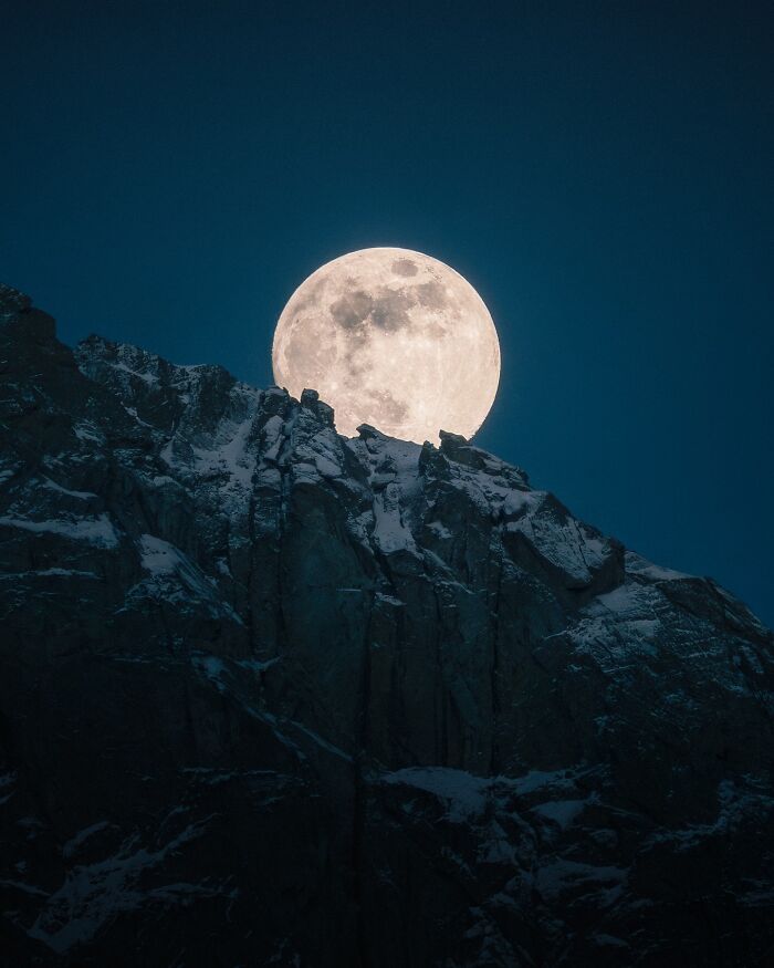 Full moon rising behind jagged snow-covered mountain peaks in a breathtaking landscape that looks straight out of a fantasy movie