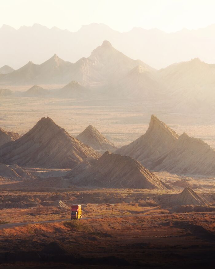 Breathtaking landscape shot featuring rugged mountains and a solitary truck on a dusty road under soft, hazy sunlight.
