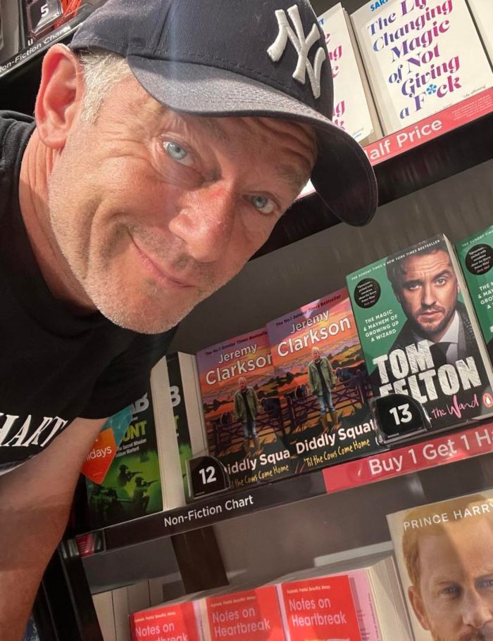Man wearing a New York Yankees cap smiles near a bookshelf featuring Tom Felton's book and Harry Potter stars reunion news.