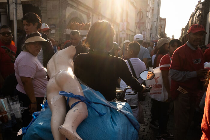 Crowded public street scene capturing a coincidental moment with a person carrying mannequin legs in golden sunlight.