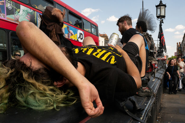 Young people relaxing on a bench near a red double-decker bus in a candid public moment captured by a photographer.