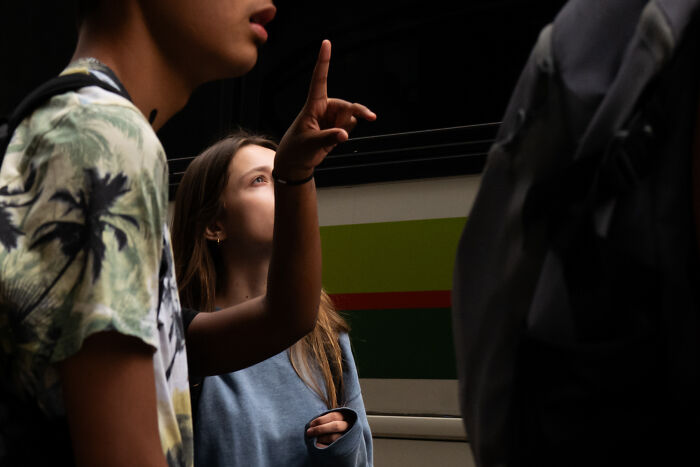 Young woman pointing while a photographer captures coincidental moments in public with people partially visible around her.