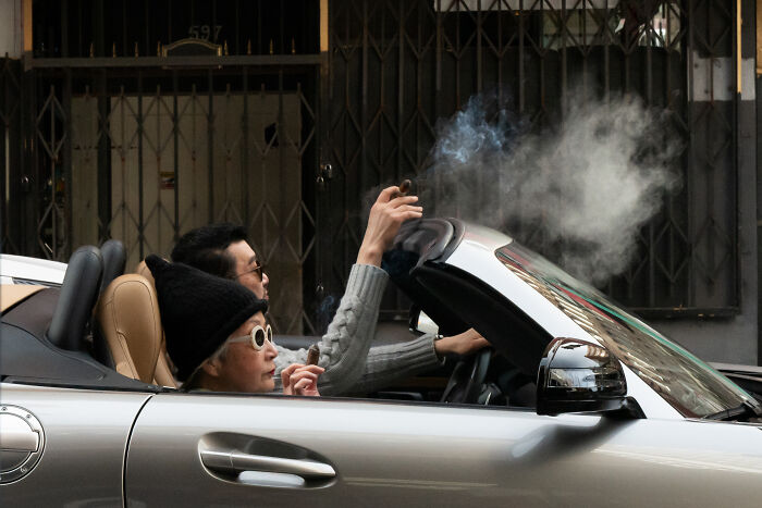 Two people smoking cigars in a convertible car, capturing a coincidental moment in public by a photographer.