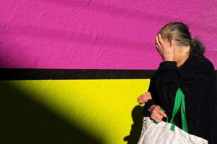 Woman covering face with hand against vibrant pink and yellow wall, capturing a coincidental moment in public by a photographer.