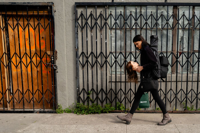 Woman carrying a realistic mannequin head walking past closed gate in a striking photographer snap of coincidental public moments.