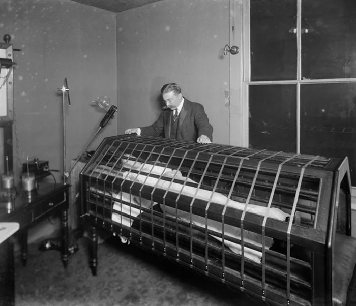 Man observes a patient inside an unsettling medical device cage, showcasing early 20th century medical equipment.