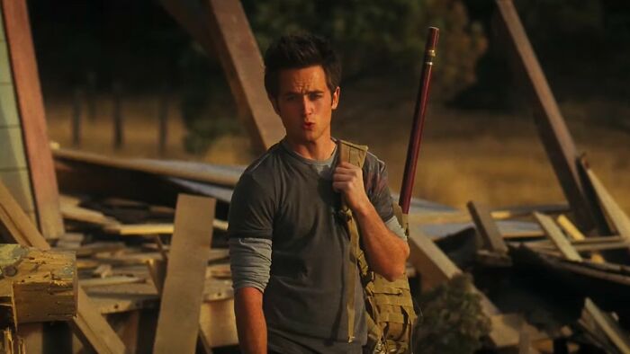 Young man in casual clothes holding a bag and staff amid ruined wooden structures in worst movies of all time scene