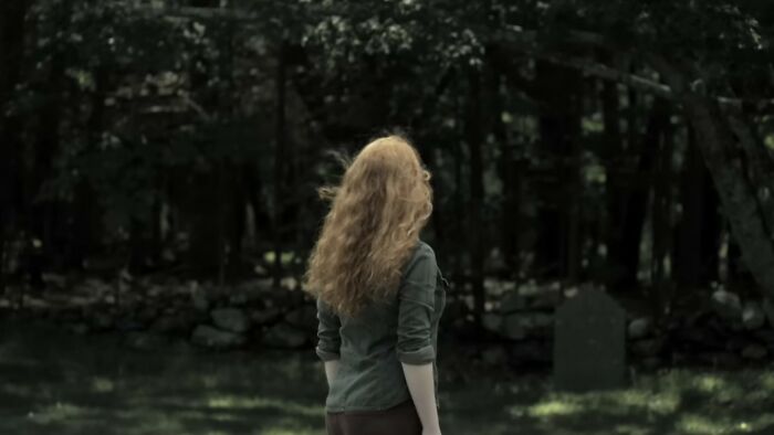 Woman with long curly hair stands in a dark forest clearing near a tombstone, evoking scenes from worst movies of all time.