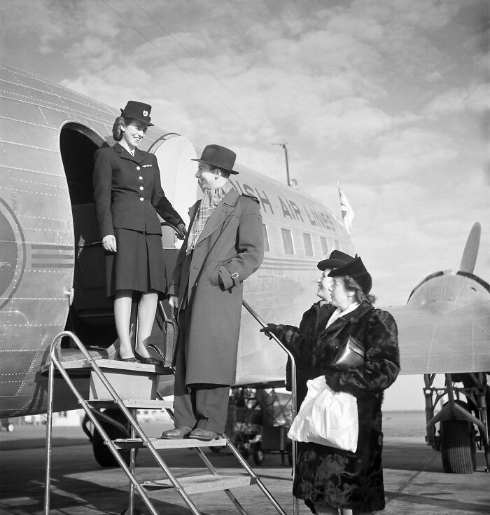 Black and white rare historical photograph of a woman flight attendant greeting passengers boarding a plane.