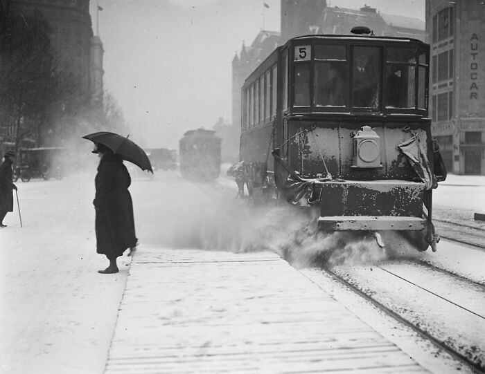 Vintage Washington, D.C. streetcar clearing snow as a person with an umbrella waits in snowy 1920s city scene.