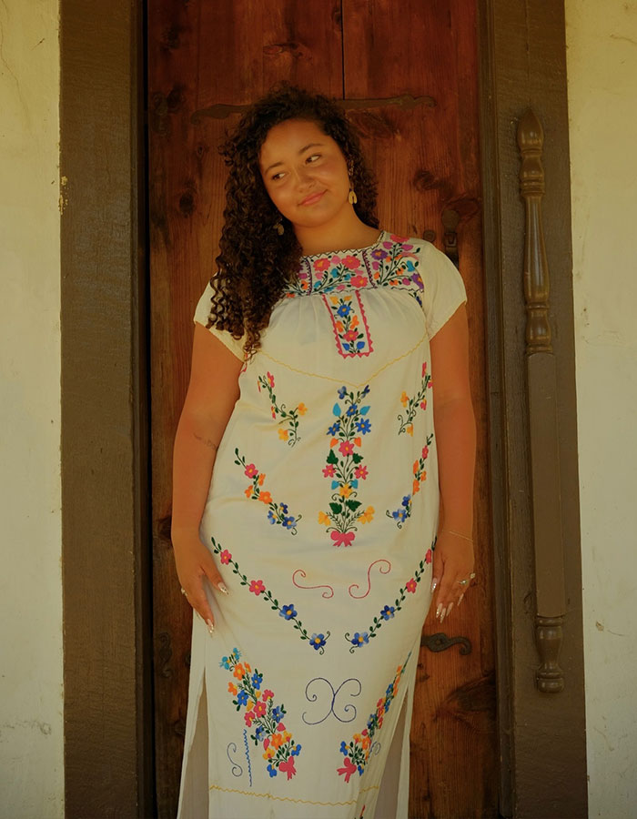 Young woman with curly hair in a floral embroidered dress standing by wooden door, related to product attorney and injury case. Young woman with curly hair in a floral embroidered dress standing by wooden door, related to product attorney and injury case.