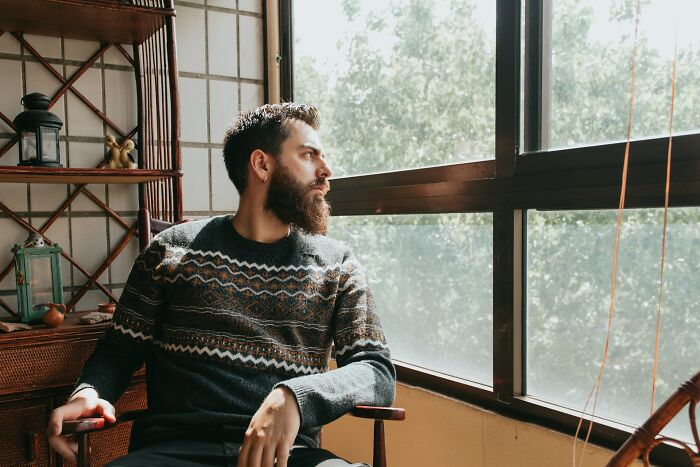 Bearded man in a patterned sweater sitting by a window, reflecting on things normal to men but bizarre to women.