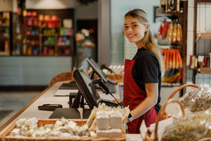 Smiling employee in a red apron standing at a checkout counter, highlighting ableist manager workplace accommodation issues. Smiling employee in a red apron standing at a checkout counter, highlighting ableist manager workplace accommodation issues.