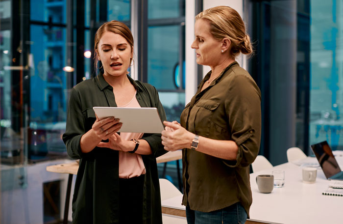 Two women in an office discussing on a tablet, depicting interaction between a manager and a disabled employee. Two women in an office discussing on a tablet, depicting interaction between a manager and a disabled employee.