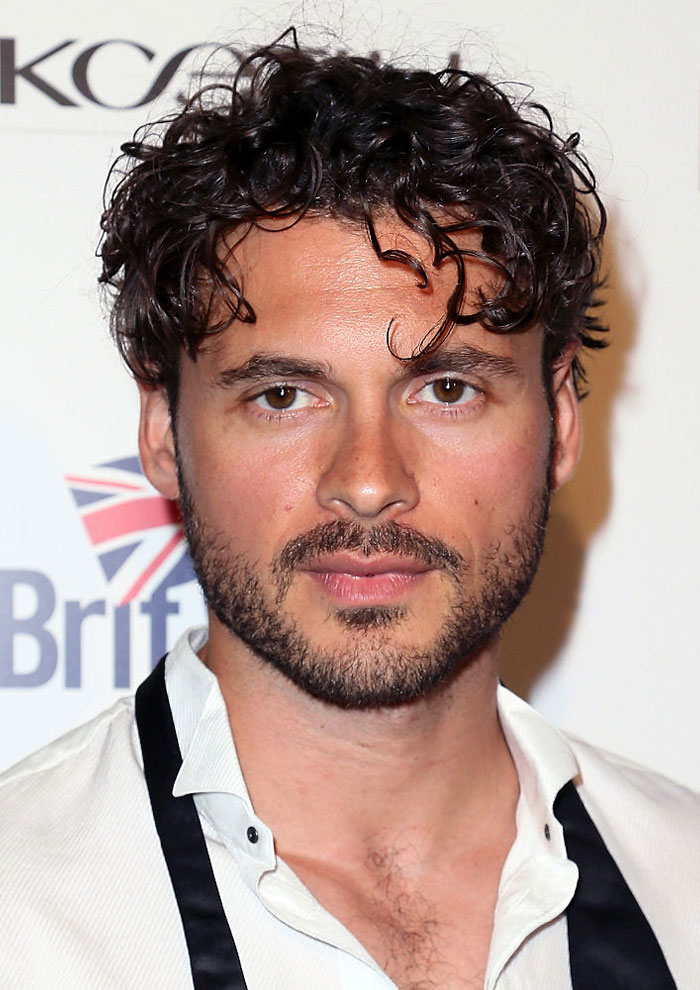 Adan Canto with curly hair and beard, wearing a white shirt, posing at an event highlighting his iconic TV roles.