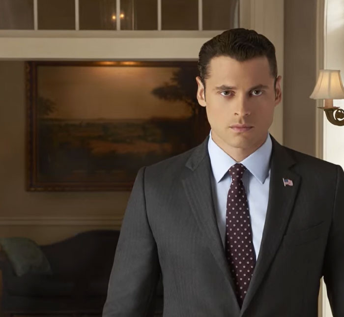 Adan Canto in a dark suit and tie standing in a formal room, representing iconic TV roles in a serious pose.