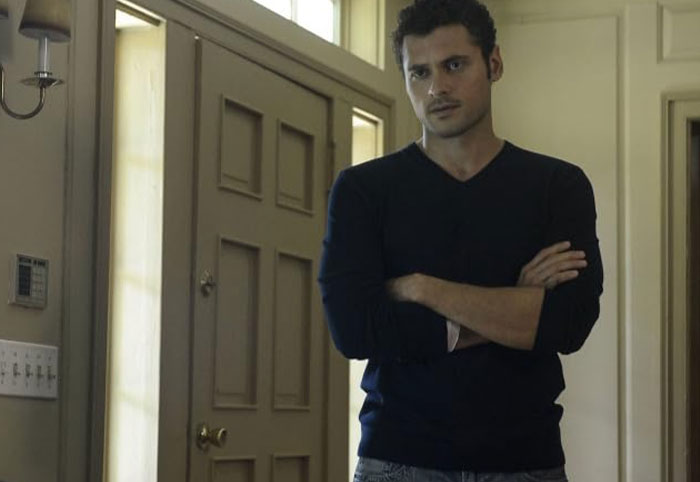 Adan Canto standing with arms crossed in a dark shirt, portraying one of his most iconic TV roles in a home setting.