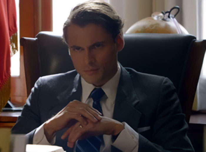 Adan Canto dressed in a suit with a blue tie, sitting thoughtfully in a dark chair in an office setting.