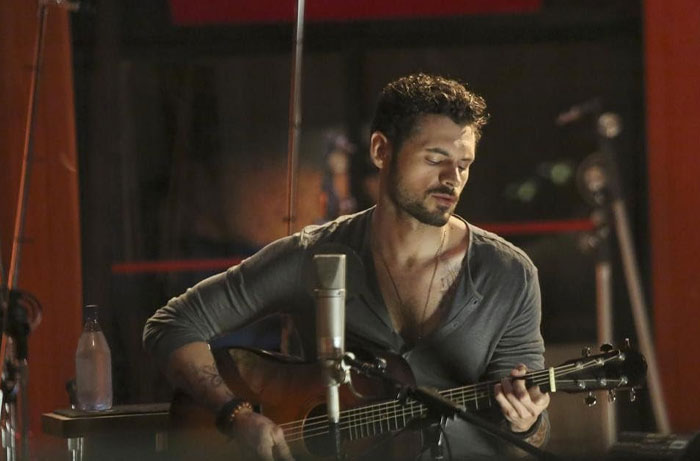 Adan Canto playing guitar in a dimly lit room, portraying one of his most iconic TV roles.