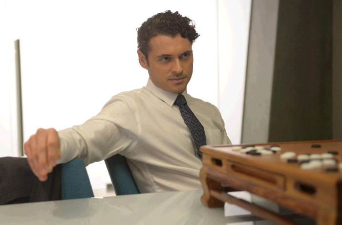 Adan Canto in a TV role, wearing a white shirt and tie, focused on a board game in a modern office setting.
