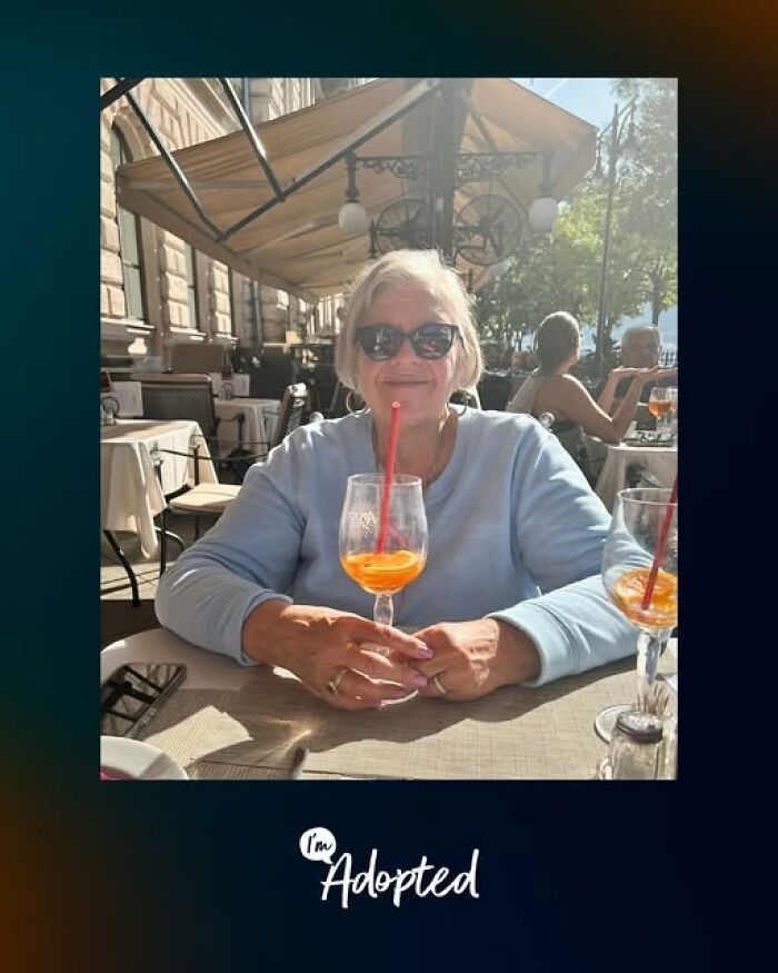Older woman wearing sunglasses, sitting at an outdoor café table with a drink, representing adoption stories.