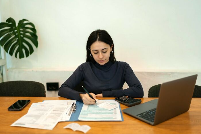 Young woman working at desk with laptop and documents, focused on writing, representing feminist daily wins concept.