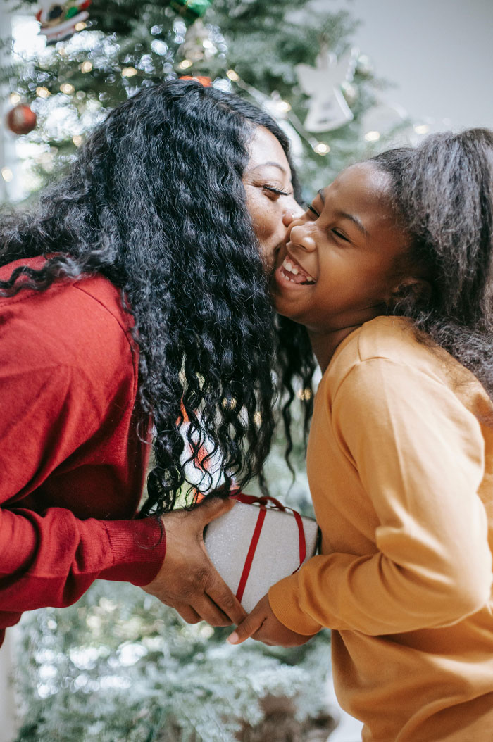 Woman kissing smiling girl while holding gift near Christmas tree, representing air signs zodiac survival kit connection.