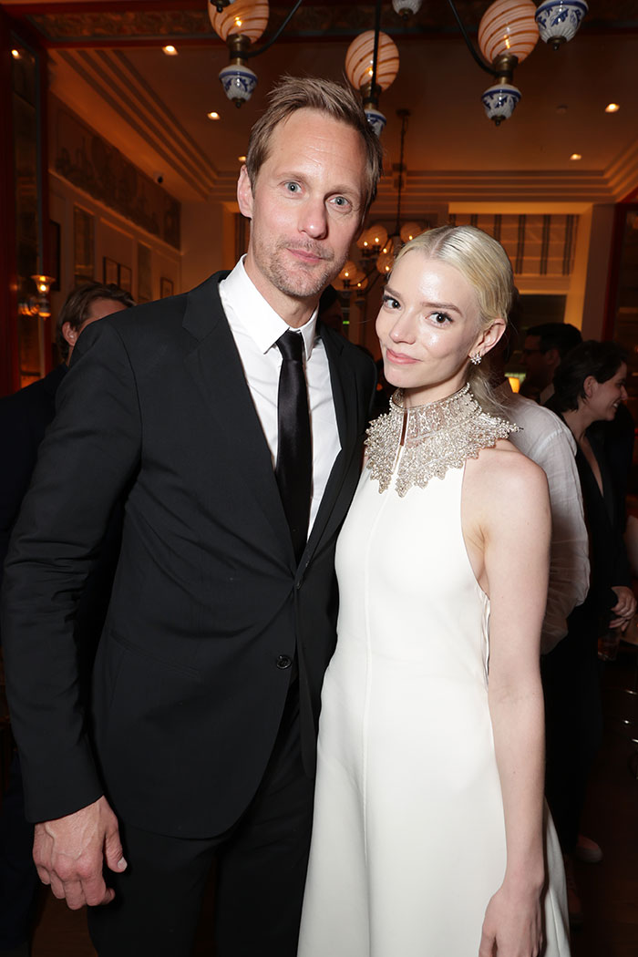 Tall male star in black suit with a shorter female star in white dress at an indoor event with warm lighting.
