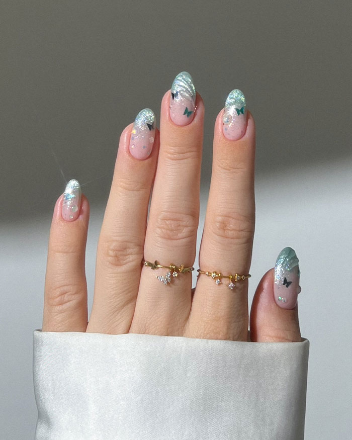 Hand with almond nails featuring silver glitter tips and butterfly decals, showcasing popular almond nails trends.
