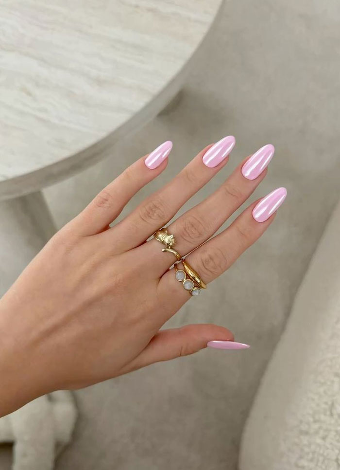 Hand with almond nails painted in glossy light pink polish, adorned with gold rings showcasing almond nails trends.
