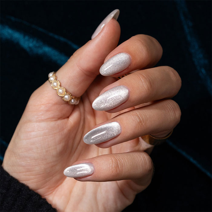 Hand with almond nails painted in shimmering silver polish, wearing pearl and gold rings against a dark background.
