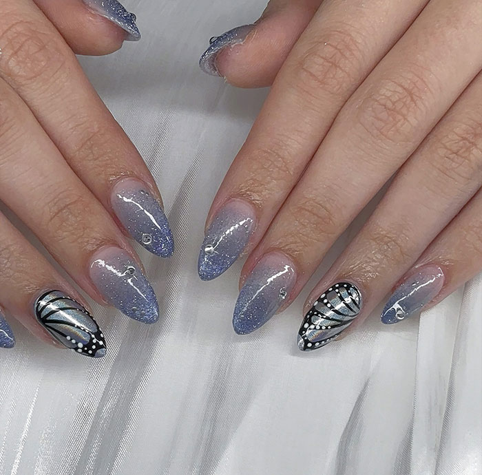 Close-up of hands with trendy almond nails featuring blue glitter ombre and black silver butterfly designs.