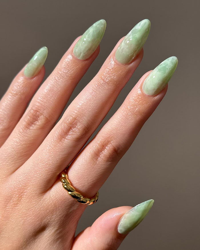 Hand with green almond nails and a gold twisted ring showcasing almond nails trends for 2025 in a neutral background.