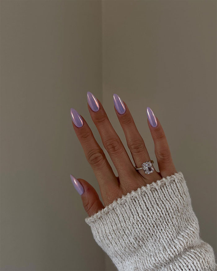 Hand with shiny purple almond nails and a large rectangular-cut diamond ring, wearing a cream knit sweater sleeve.