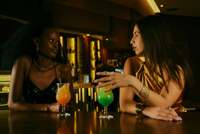 Two women discussing outrageous student names while seated at a bar with colorful cocktails in a dimly lit setting.
