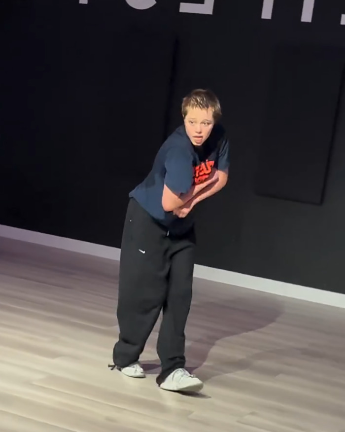 Shiloh Jolie-Pitt, 19, wearing casual clothes, performing a dance move in a studio with hardwood floors and dark walls.